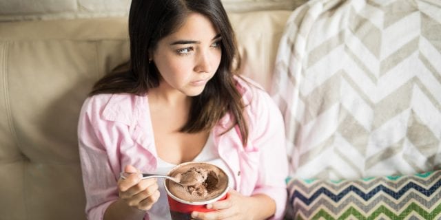 A woman in a pink shirt is sitting on a couch eating out of a carton of ice cream while looking to the side with a sad expression.