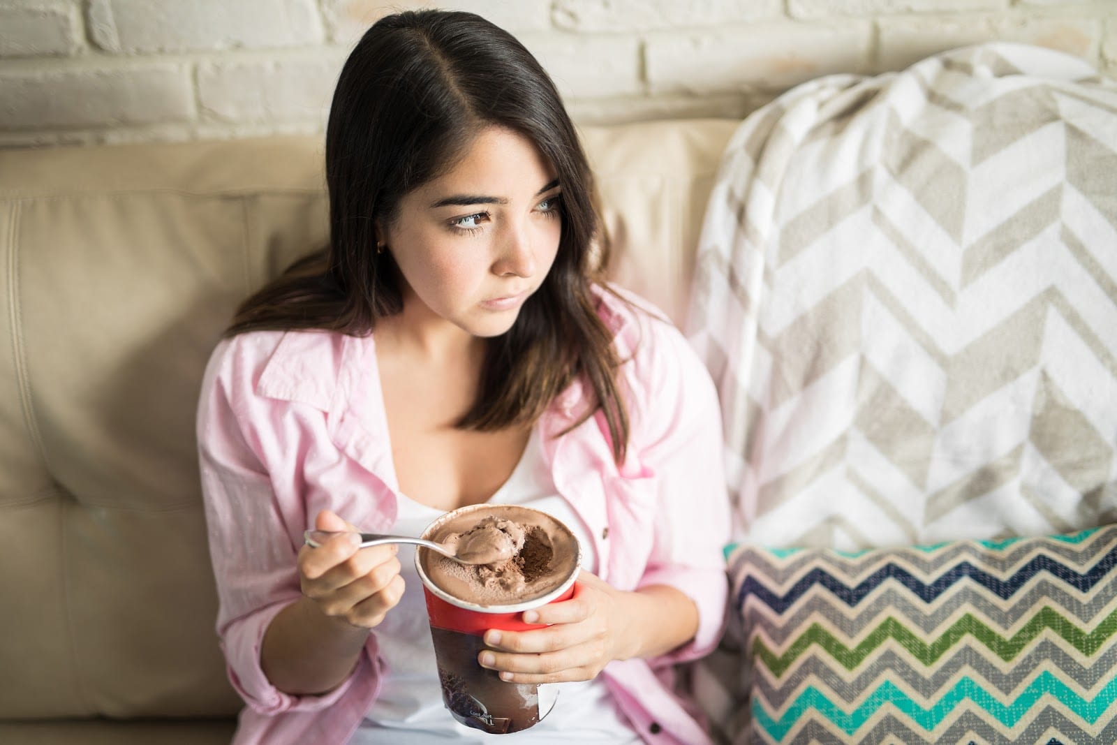 A woman in a pink shirt is sitting on a couch eating out of a carton of ice cream while looking to the side with a sad expression.