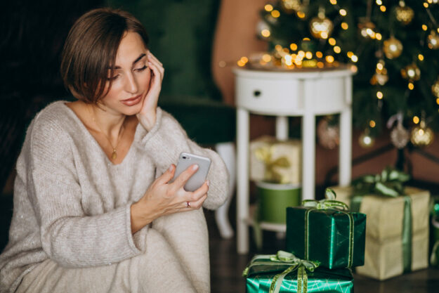 Woman sitting by Christmas tree and shopping on the phone