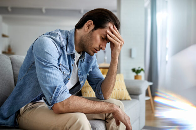 Young man holding his head in pain while sitting on the sofa.