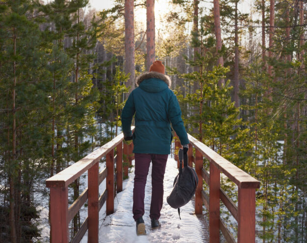 Walking in the forest. Man in green jacket and orange hat go to the sun with backpack in one hand across mountain river bridge. Travel concept with healthy life and fresh air.